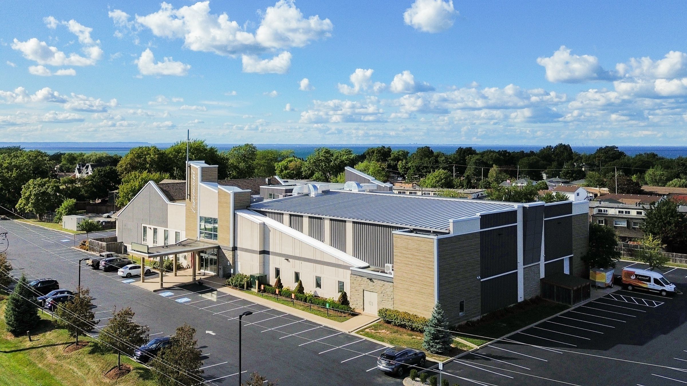 An aerial view of Lakemount Worship Centre, a long modern building of grey and buff panels with a tall entrance tower and a canopy over the doors, its parking lot around it and Lake Ontario on the horizon