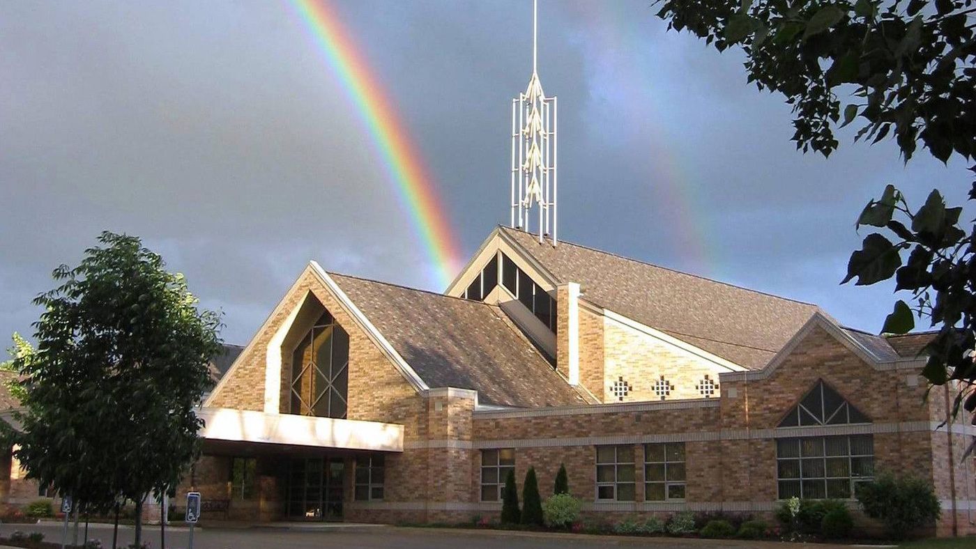 Mountainview Christian Reformed Church in evening sun under a stormy sky with a rainbow, a buff-brick church with steep glazed gables, an open steel spire and a canopy over the entrance