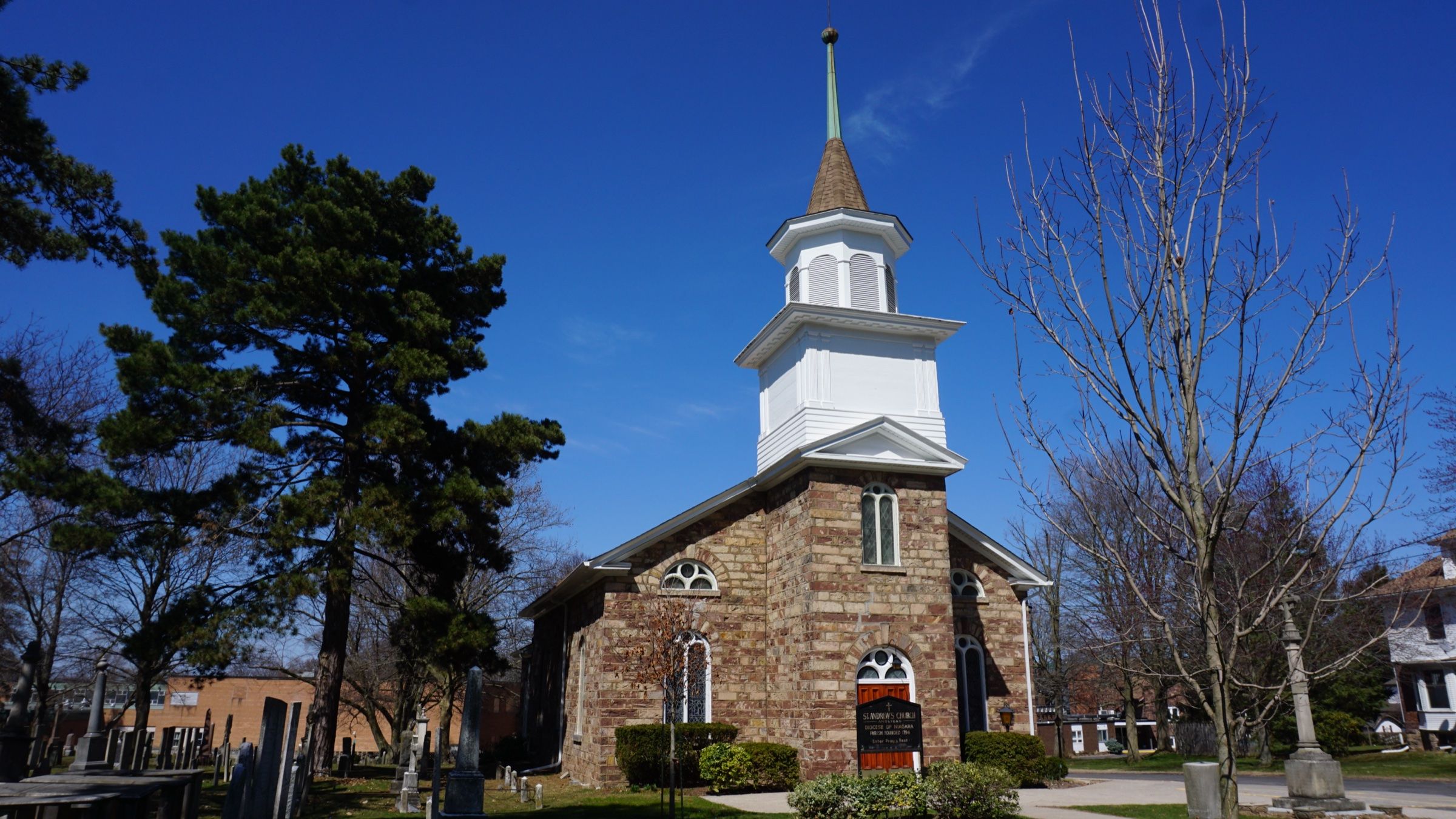 The stone front and white wooden bell tower of St. Andrew's Anglican Church, with old gravestones in the churchyard beside it