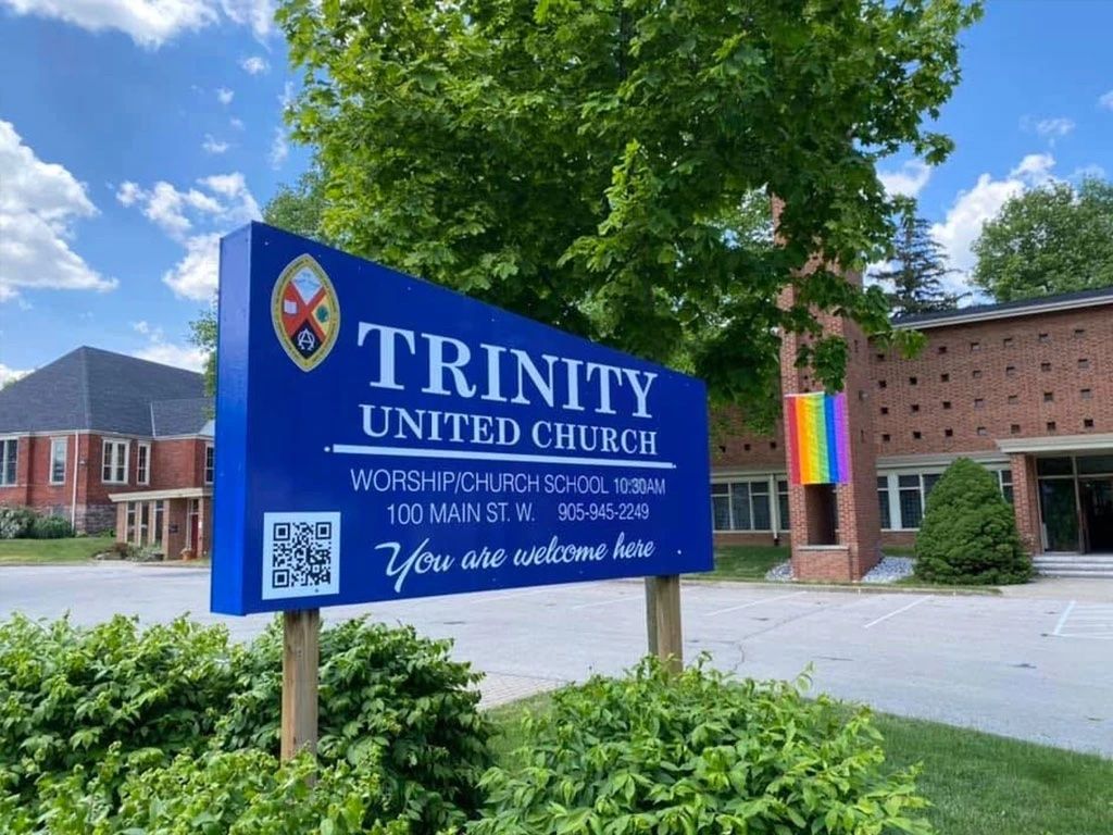 Trinity United Church's blue roadside sign on Main Street West, with the church's brick bell tower and entrance behind it