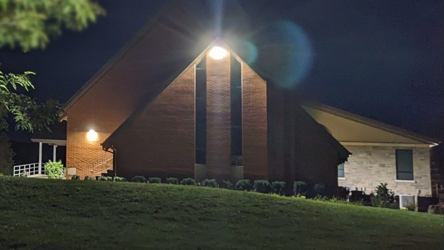 Vineyard Canadian Reformed Church in Beamsville at night, its tall brick gable lit by a lamp at the peak between two narrow windows, with a stone-clad wing beside it